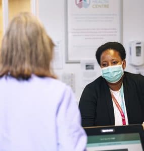 A GP receptionist looking at a patient at the reception desk of a GP surgery.