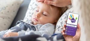 بستر پر ایک بچہ، اپنے سر پر ہاتھ رکھ کر اپنا درجہ حرارت چیک کر رہا ہے۔ تصویر کے کونے میں اسمارٹ فون پکڑے ہوئے ہاتھ میں ہے، فون کی اسکرین پر HANDi ایپ ہے۔