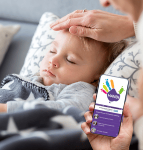 A child in a bed, with a hand on their head checking their temprature. A hand holding a smartphone is in the corner of the image, the phone has the HANDi app on the screen.