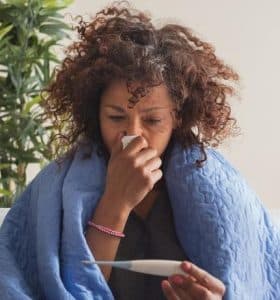 Woman wrapped in a blue blanket, blowing her nose with a tissue and looking at a thermometer.