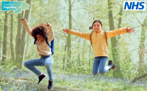Two children smiling and jumping in a forest. The Healthier Together logo is in the top left corner and the NHS lozenge logo is in the right hand corner.